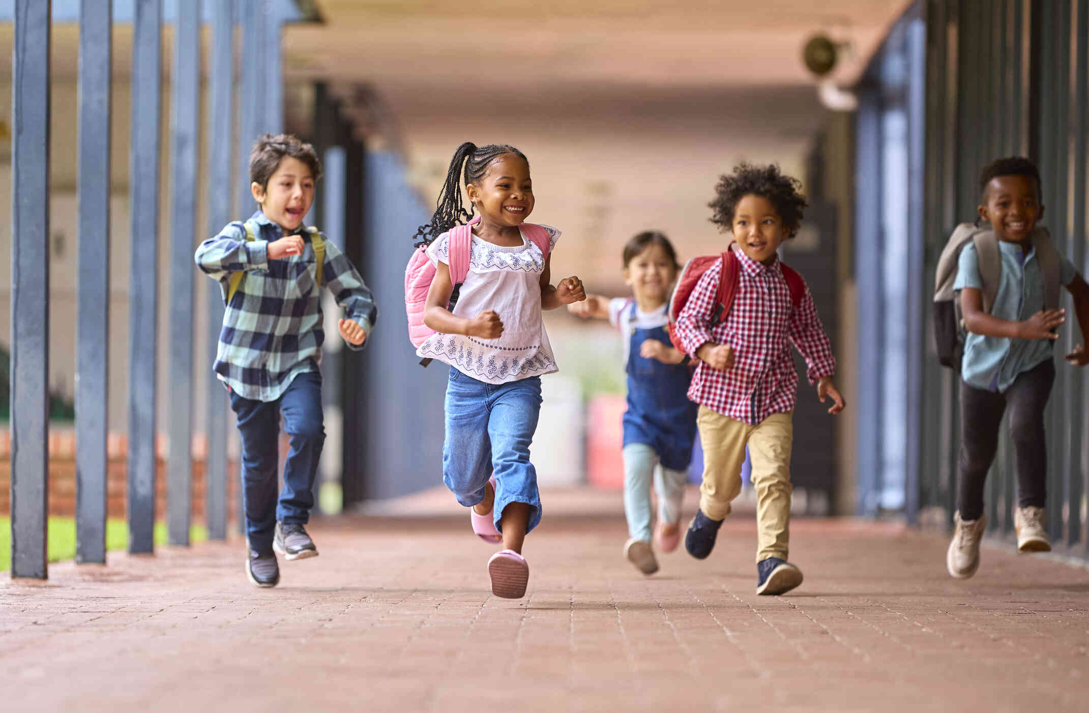 A group of kids wearing backpacks run down a breezeway of a school while smiling.